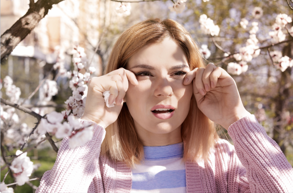 A young woman standing in front of a flower tree wiping her eyes because of her allergies. 