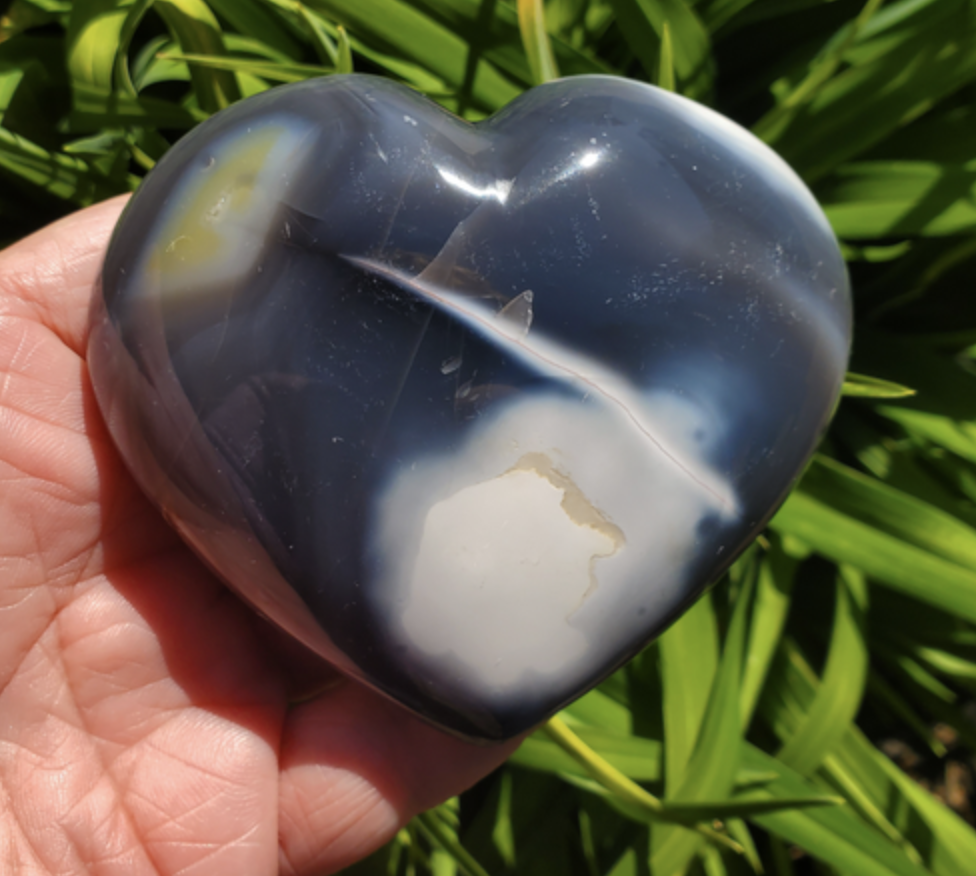 A large blue lace agate heart crystal in a woman's hand with plants in the background.