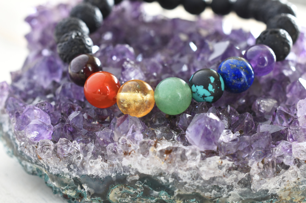 A chakra bracelet with multiple round stones of different colors sitting on a chunk of purple amethyst. 